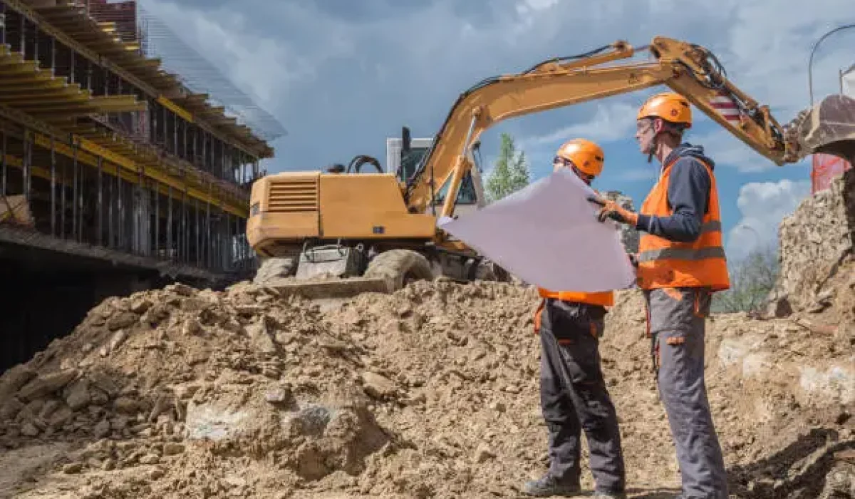 Deux ouvriers en sécurité sur un chantier, examinant un plan devant une excavatrice et un bâtiment en construction. agenci intérim bâtiment industrie